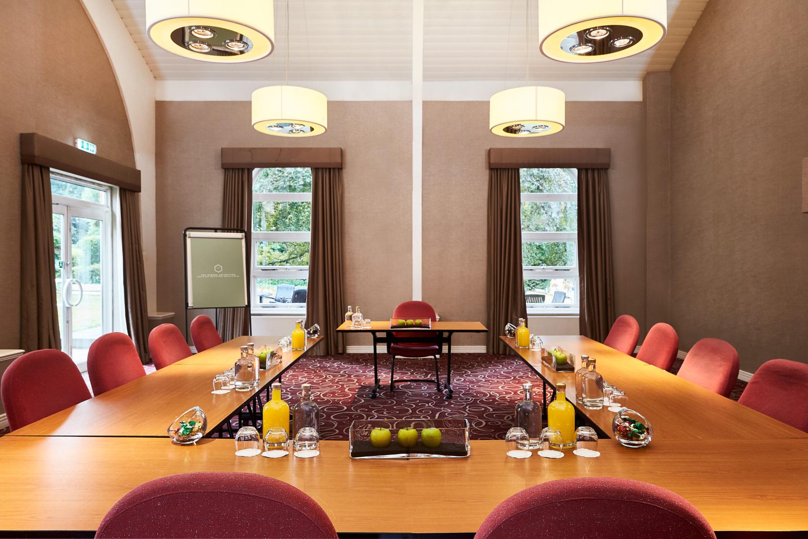 Conference room with red chairs, wooden tables set with glasses, glass water, a juice bottle, green apples, and sweets in a glass bowl.