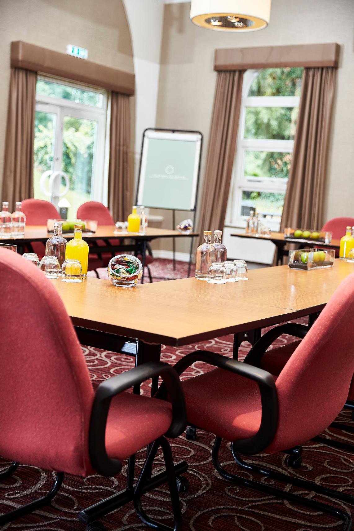 Conference room with red chairs, wooden tables set with glasses, glass water, a juice bottle, green apples, and sweets in a glass bowl.